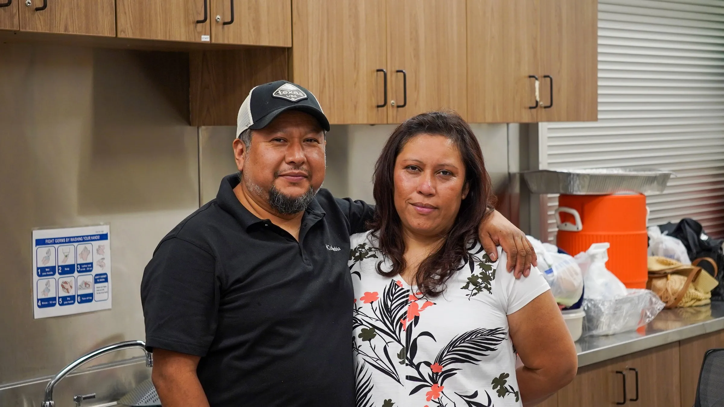 A couple stands arm in arm in a community hall kitchen
