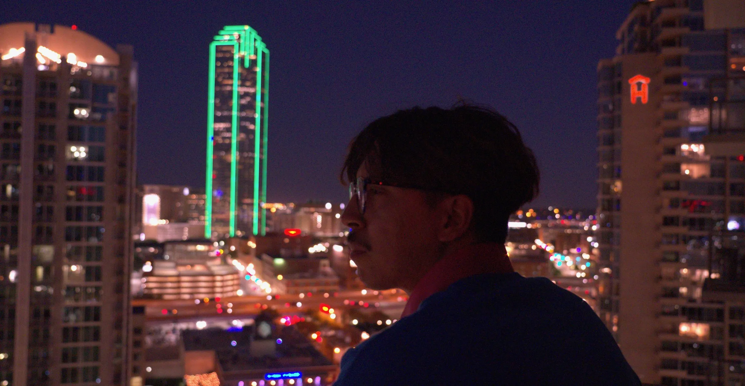 A man in silhouette against the Dallas skyline at night, the green-lit Bank of America Plaza behind him
